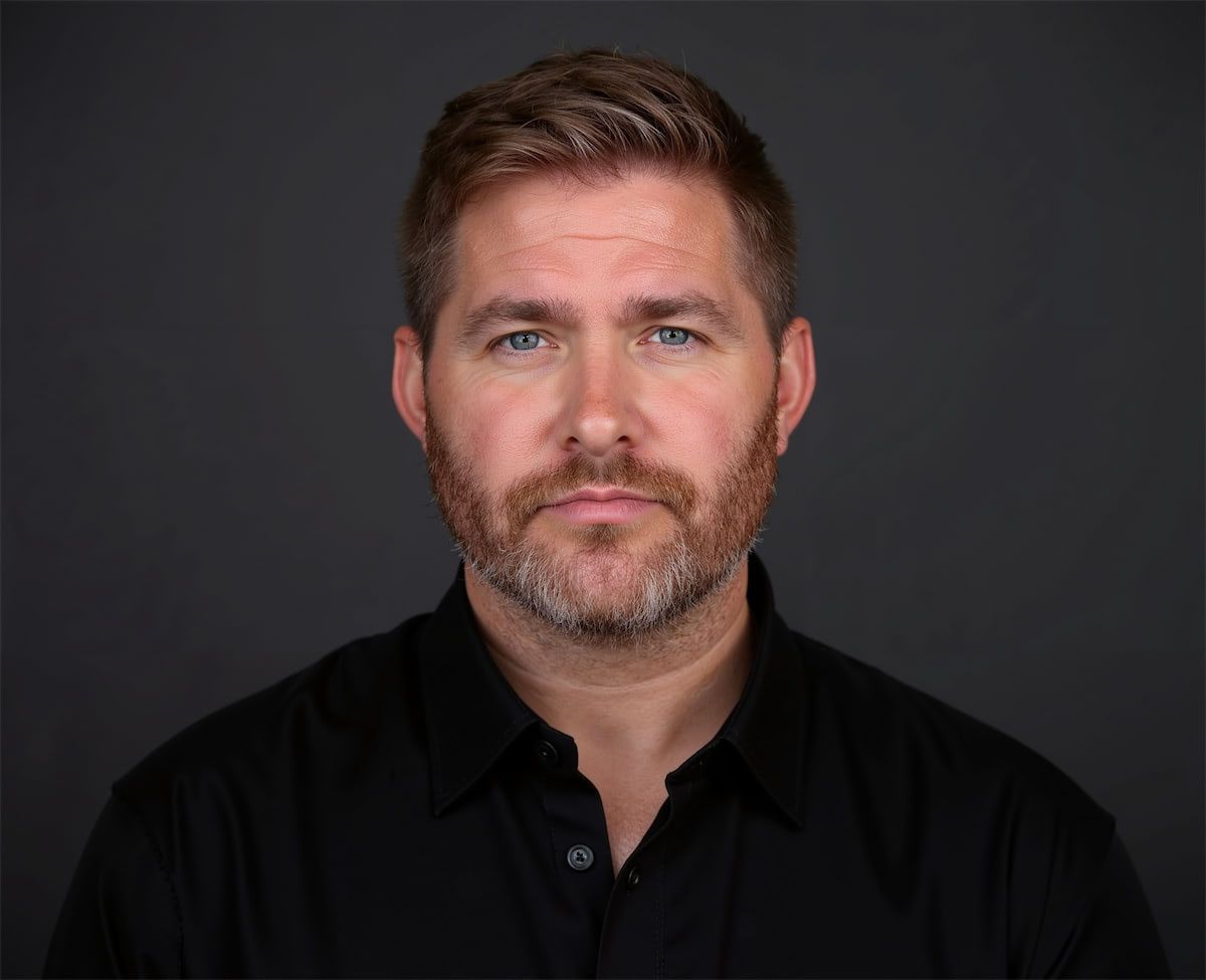 A headshot of a person with short brown hair and facial hair, wearing a black collared shirt against a dark background.