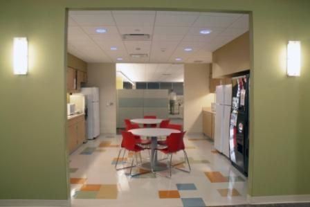 An office breakroom with two round tables, red chairs, refrigerators, and a vending machine on a multicolored tiled floor.