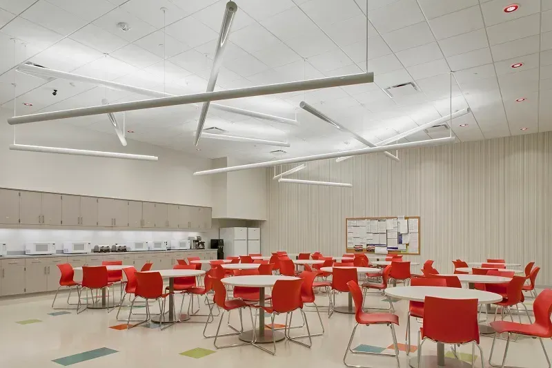 A break room with round tables and bright orange chairs, featuring beige cabinets and modern overhead lighting.