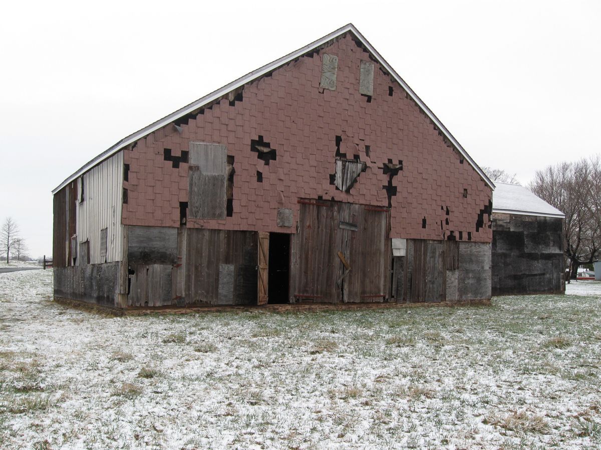 A weathered, red-sided barn with missing boards stands in a field lightly dusted with snow under an overcast sky.