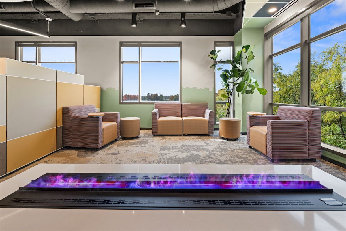 Modern office lounge with beige chairs, large windows, plants, and a purple-lit linear fireplace or water feature
