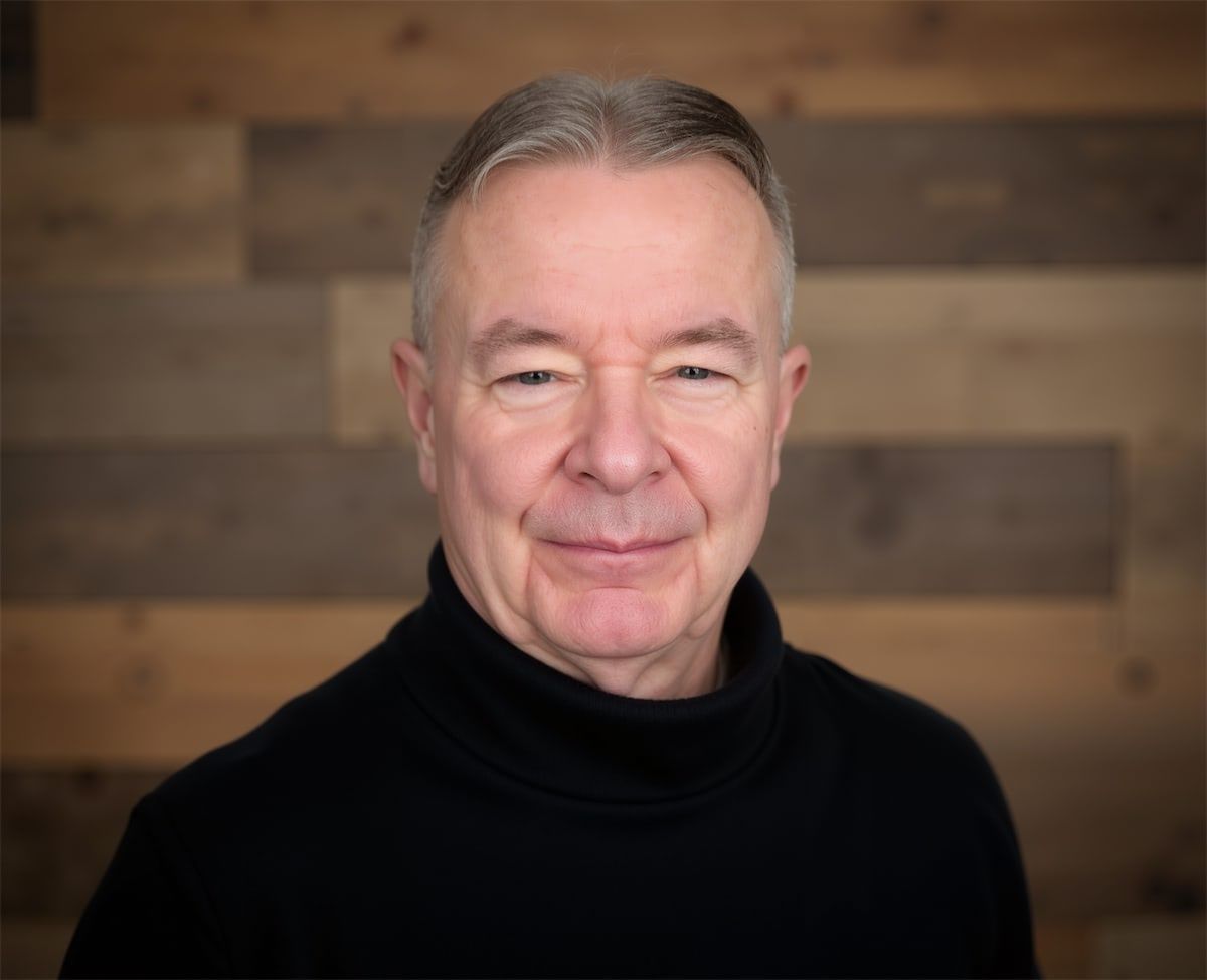 A person wearing a black turtleneck stands in front of a brown, horizontally-planked wooden wall.