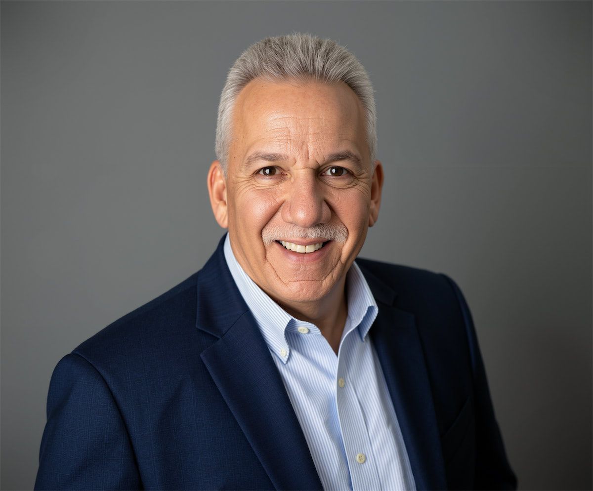 Headshot of a smiling person with short grey hair, wearing a light blue dress shirt and navy blazer against a grey backdrop.
