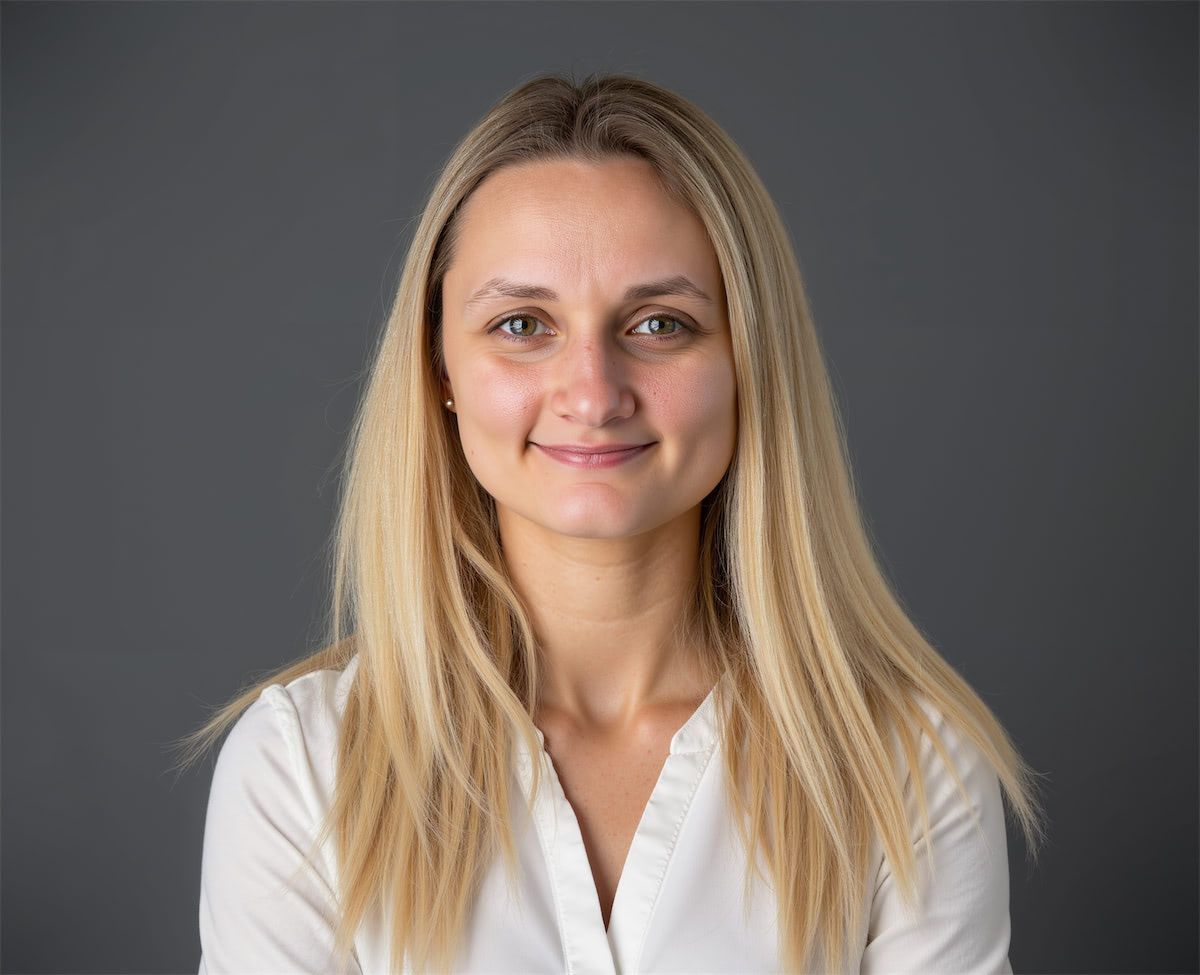 A person with long blonde hair and a neutral expression, wearing a white shirt against a plain dark gray background.