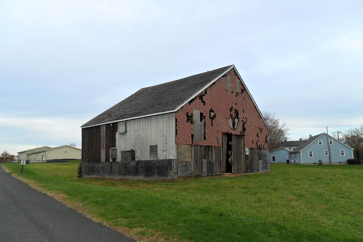 A weathered, multi-textured barn with red brick and wooden siding sits in a grassy field next to a rural road.