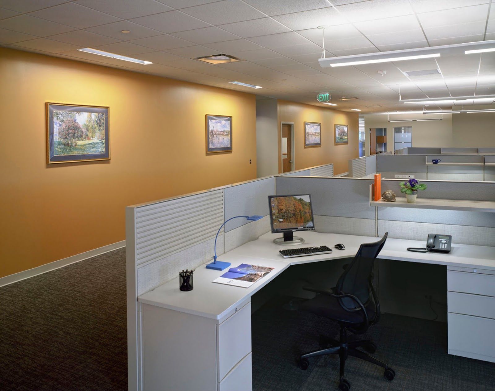 An empty office cubicle with a desk, computer, and chair, next to a hallway with orange walls and framed artwork.