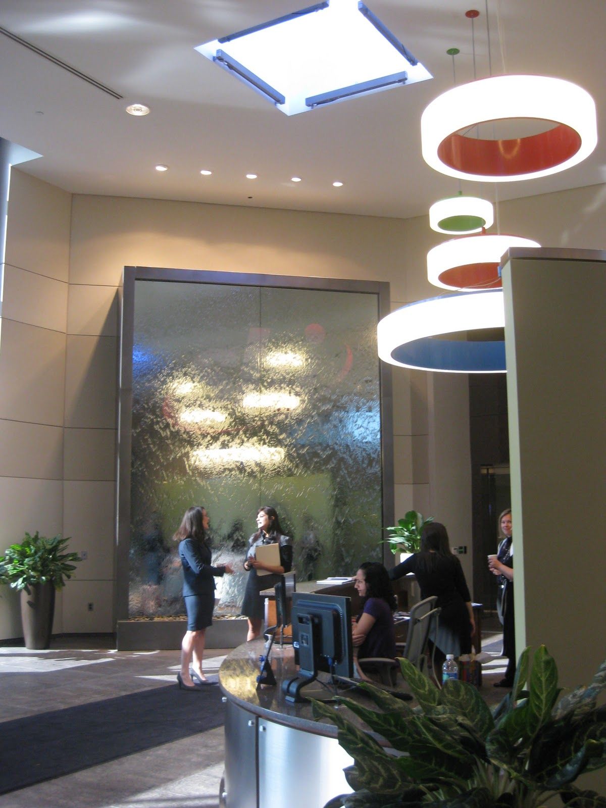 Modern lobby with a large indoor water feature, reception desk, and people standing in a bright, open space.