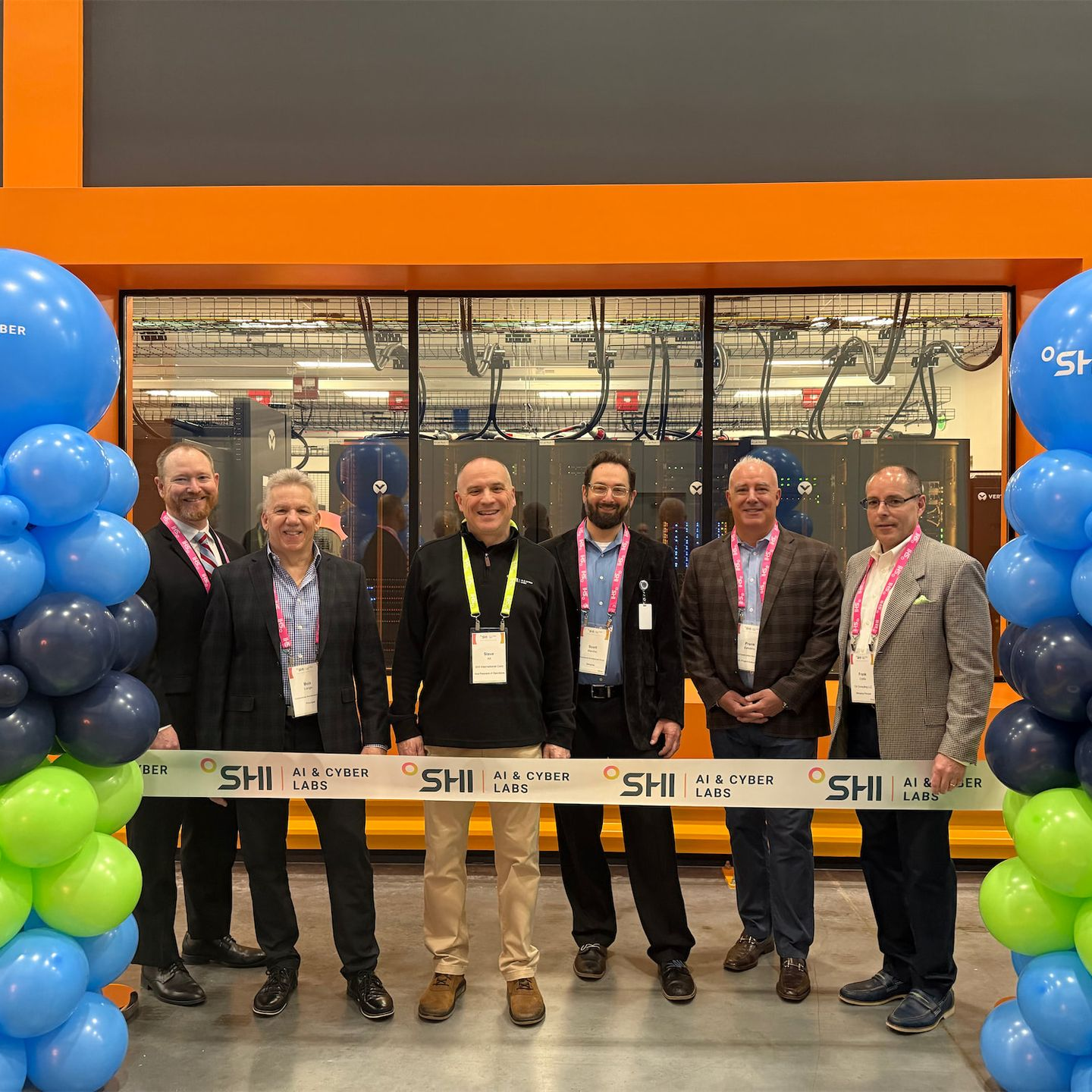 Six people in business attire stand smiling behind a ceremonial ribbon in front of a facility with large servers.