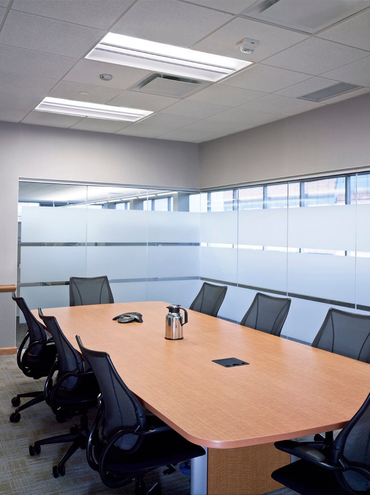 A light-wood conference table with six chairs in an office with frosted glass walls and fluorescent ceiling lights.