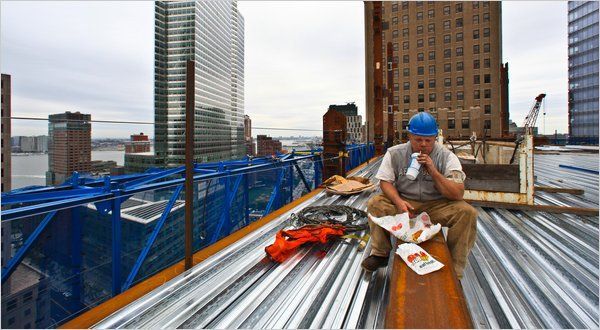 A worker in a hard hat sits on a steel beam high above a city, eating lunch on a partially constructed building.