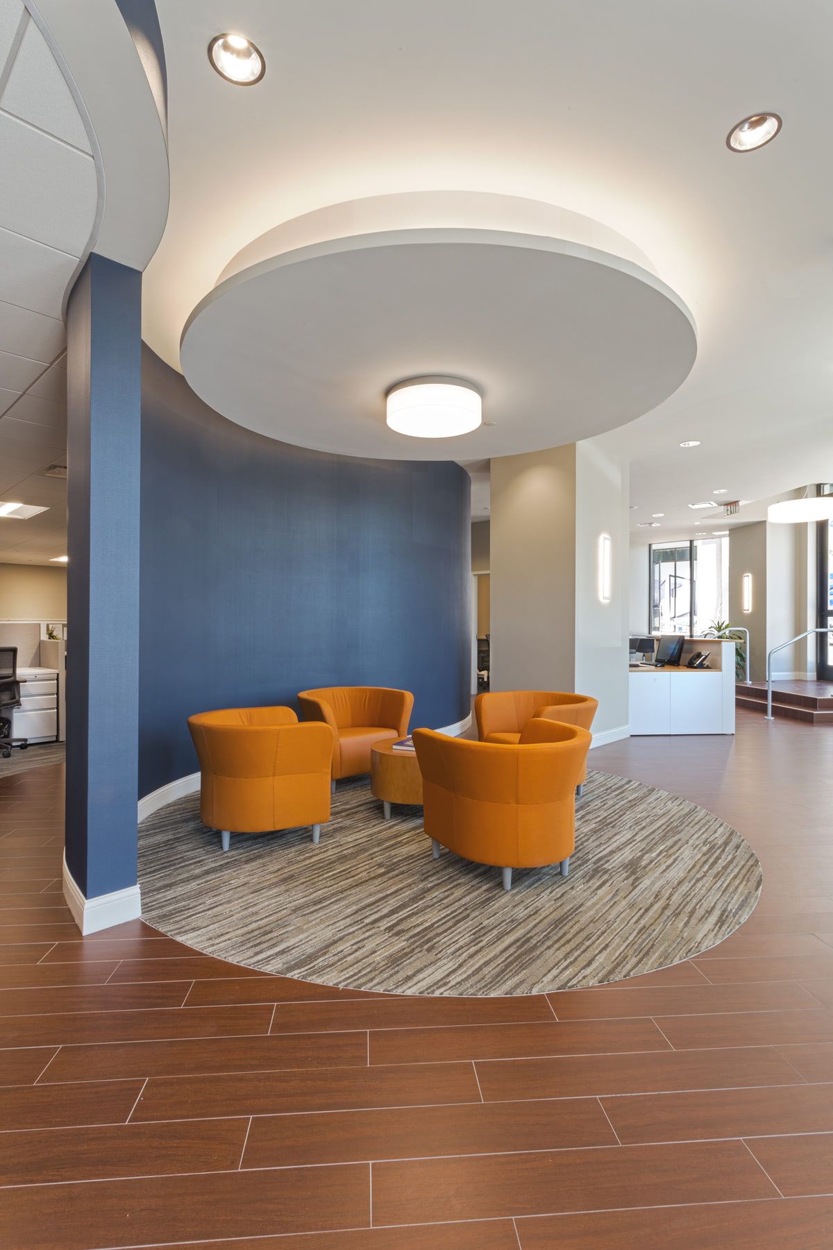 Modern office lobby with three orange chairs on a circular patterned rug in front of a curved blue wall.