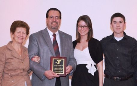 Four people pose smiling for a photo; the center individual holds an award plaque with a YMCA logo against a plain wall.