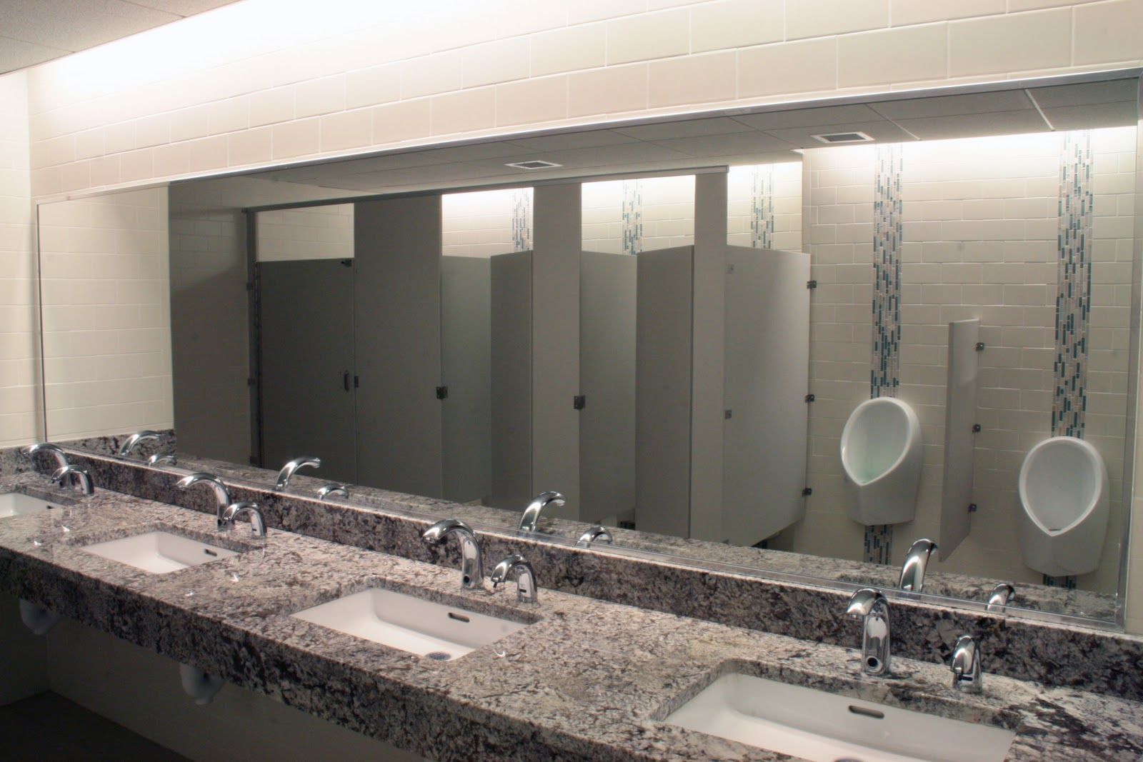 A modern public restroom with granite countertops, multiple sinks, a large mirror, and wall-mounted urinals with tile.