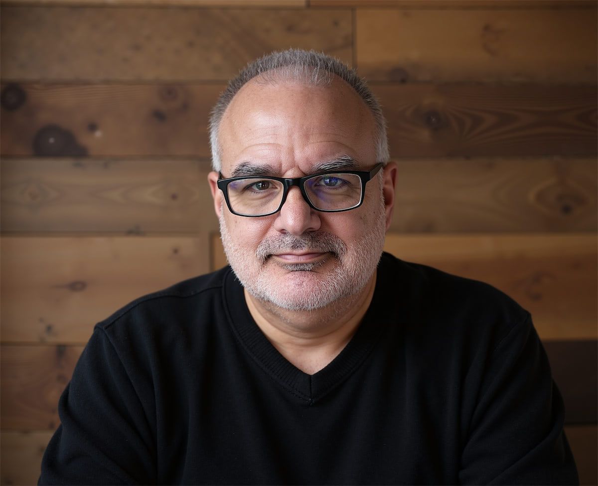 A person with short, greying hair and glasses wearing a black V-neck shirt, posing against a wooden plank wall.