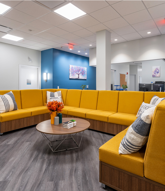 A waiting area with a yellow sectional sofa, a round coffee table with flowers, and gray flooring in a modern room.