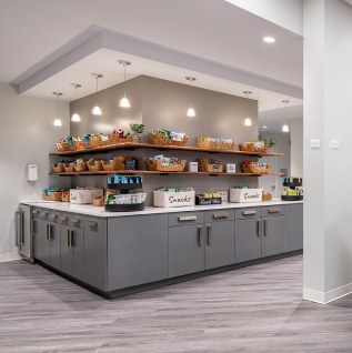 A snack station with grey cabinets, white countertops, and two wooden shelves filled with baskets of snacks.