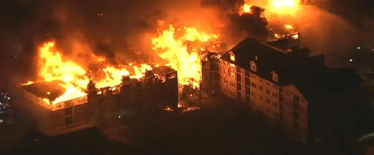 An aerial view of a multi-story building engulfed in large orange flames and smoke at night.