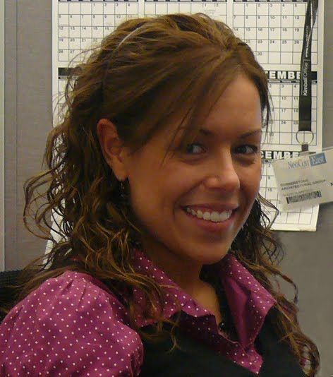 A smiling person with wavy brown hair wearing a purple, white-polka-dot shirt in front of a wall calendar.