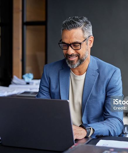 A smiling professional in a blue blazer and glasses works on a laptop in a modern office.
