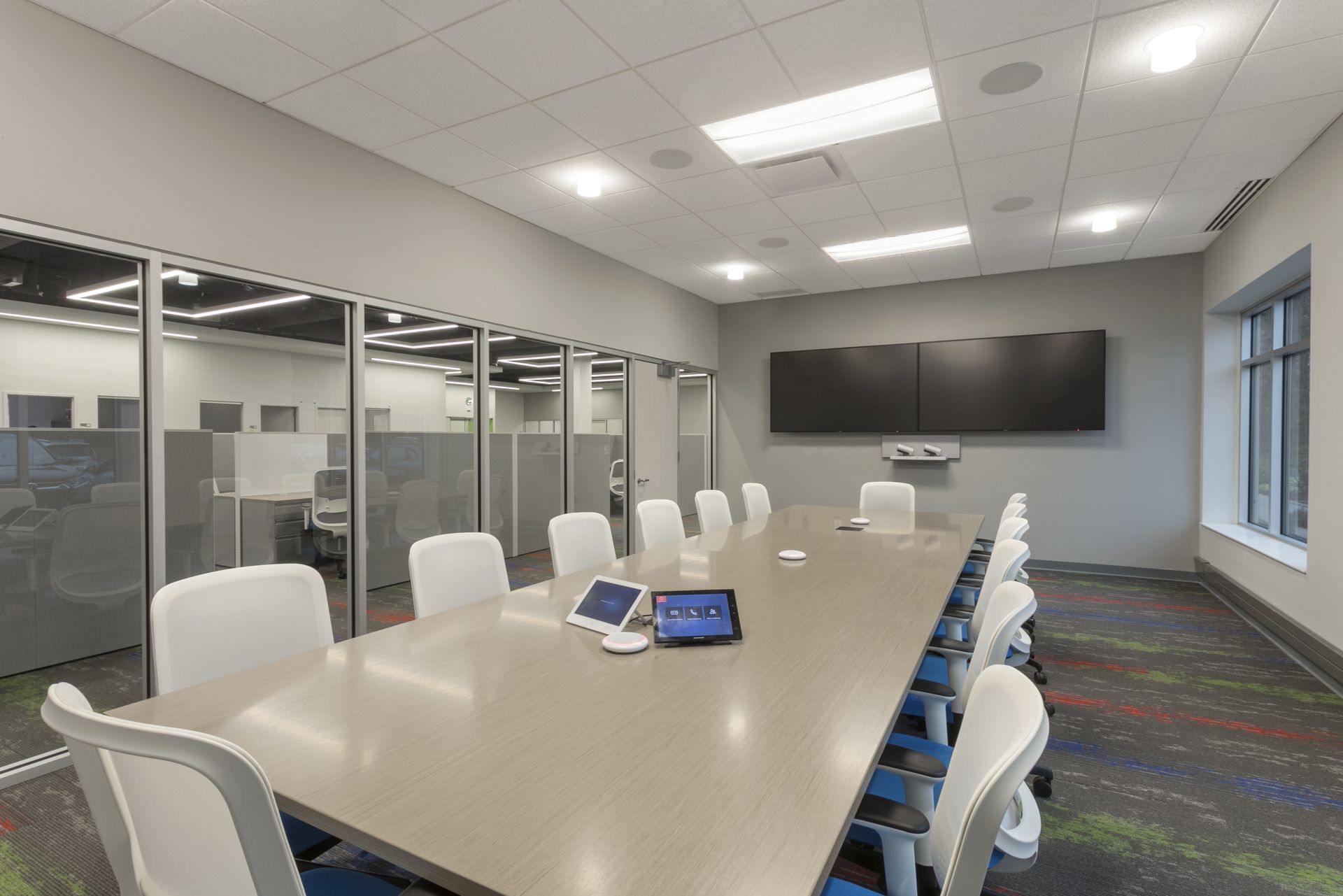 Modern meeting room featuring a long conference table, white chairs, glass walls, and a large display screen on the wall.