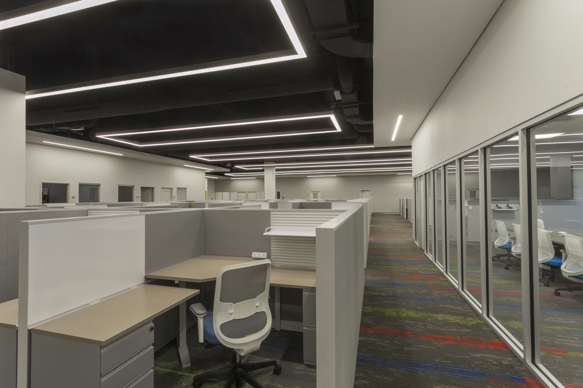 Modern office space with cubicles, gray chairs, patterned carpet, and linear ceiling lights over a glass-walled room.