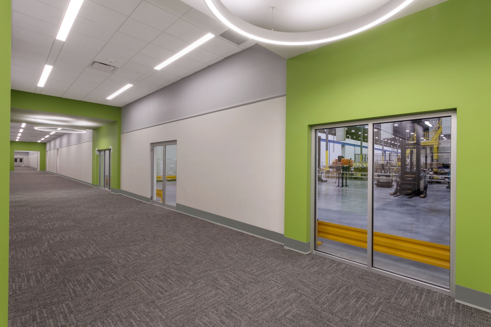 A long, carpeted hallway with lime green walls and large glass doors looking into a brightly lit industrial workspace.