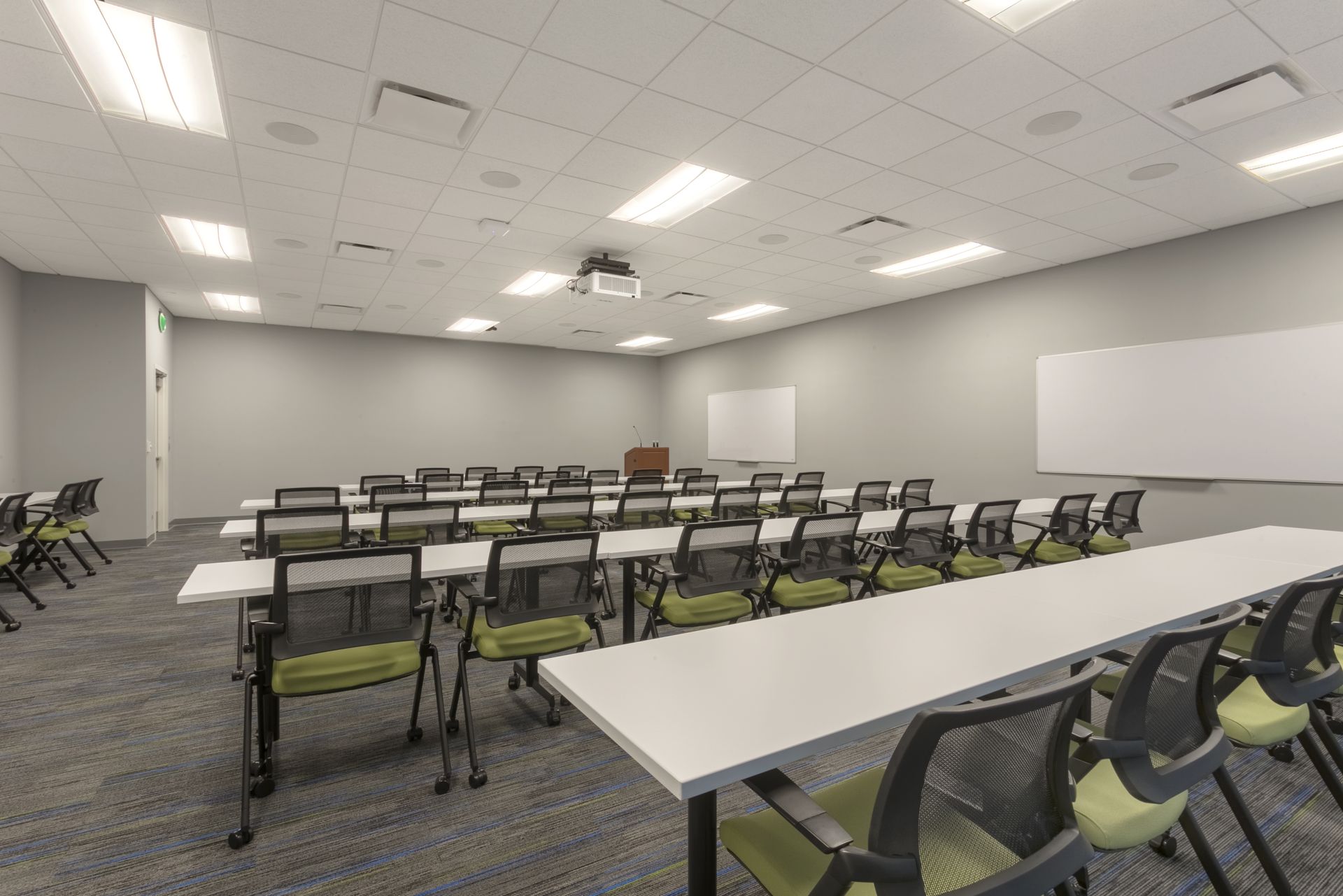 A conference room with rows of tables and chairs, carpeted floors, fluorescent lighting, and whiteboards on the walls.