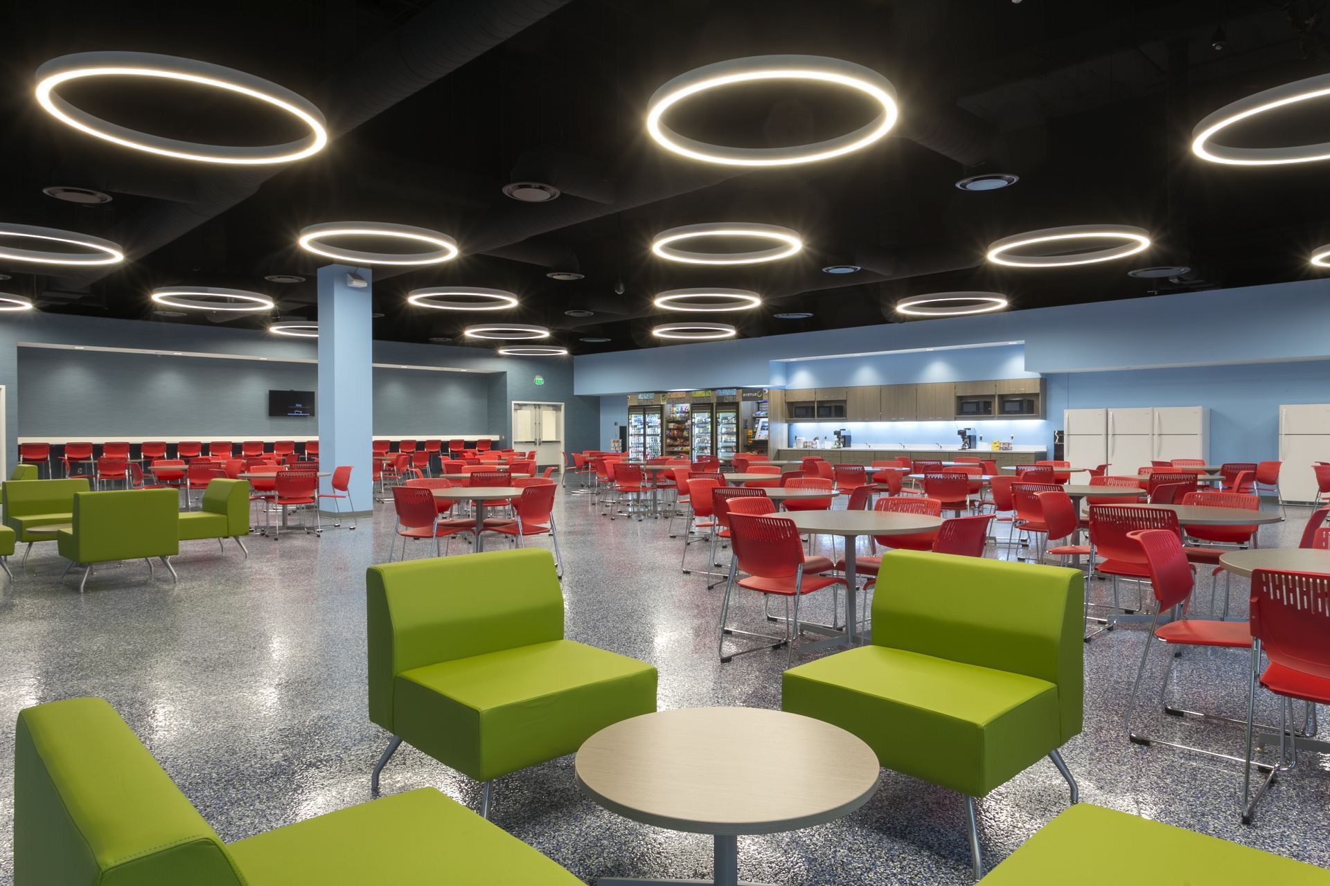 A modern, brightly lit cafeteria with green lounge chairs, red dining chairs, round tables, and circular pendant lights.