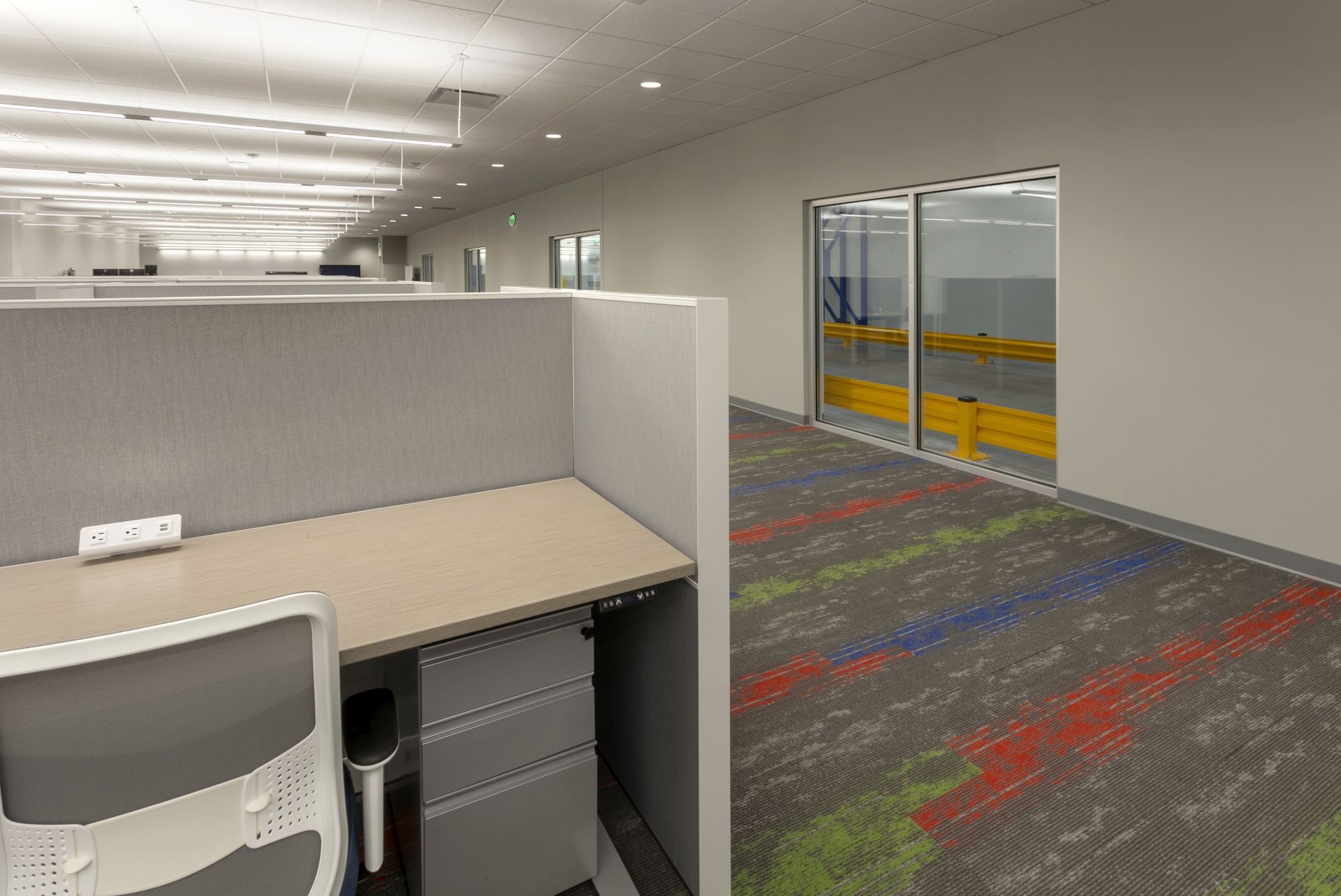 A modern office cubicle with a desk and chair next to a glass wall looking into a warehouse with yellow guardrails.