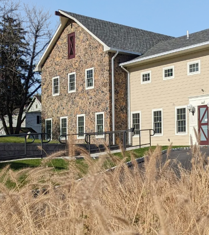 A stone barn building attached to a modern tan building behind tall, dry grass on a sunny day.
