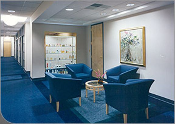 A waiting area with four blue armchairs arranged around a round glass table on a blue carpet in a brightly lit hallway.