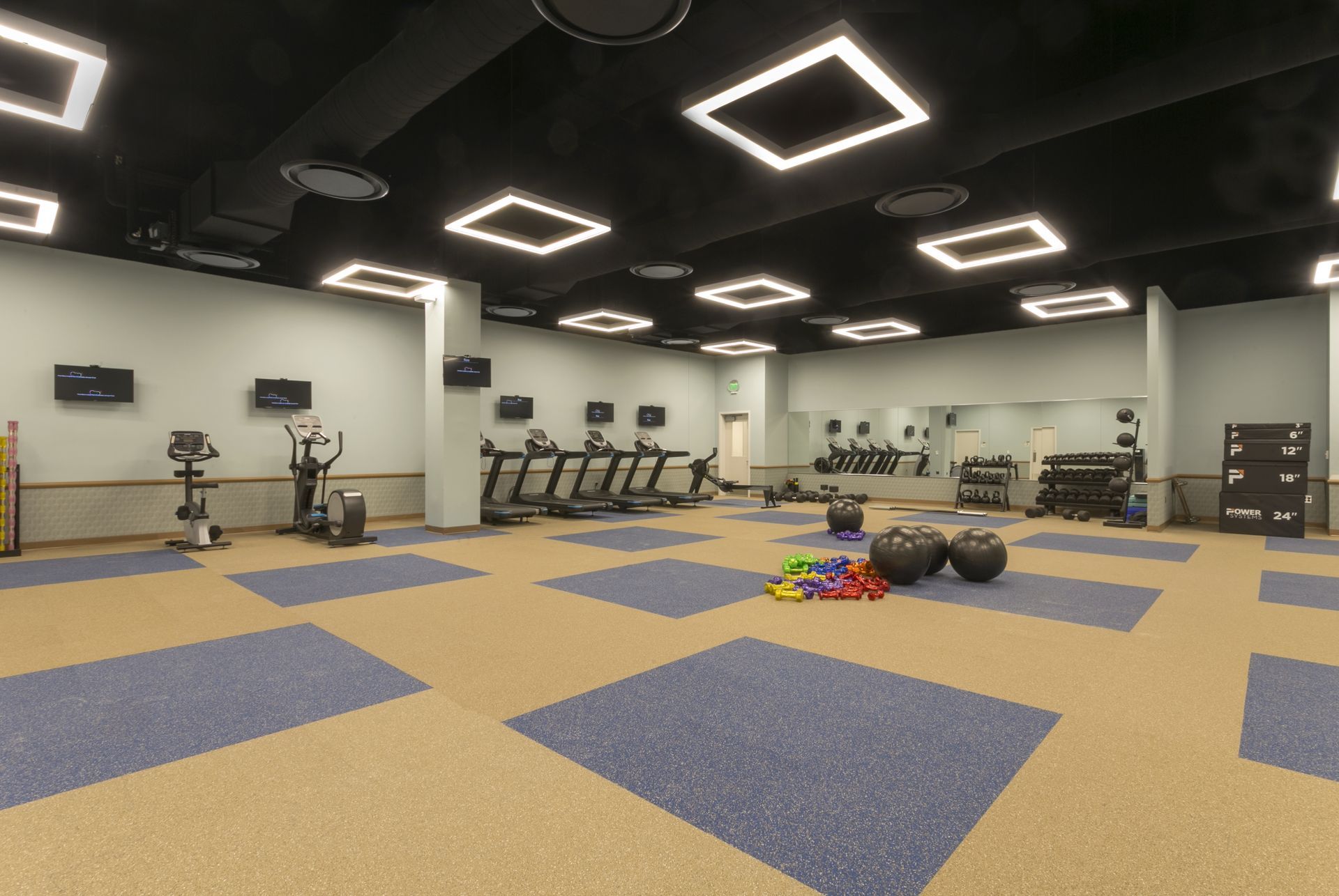 A gym with fitness equipment, treadmills, blue mat squares on a tan floor, and square overhead lights.
