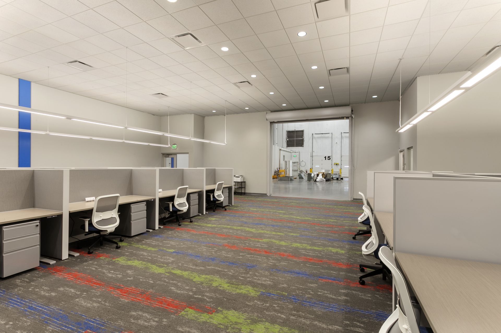 A bright, modern office space featuring rows of grey cubicles with white chairs on a patterned, multi-colored carpet.