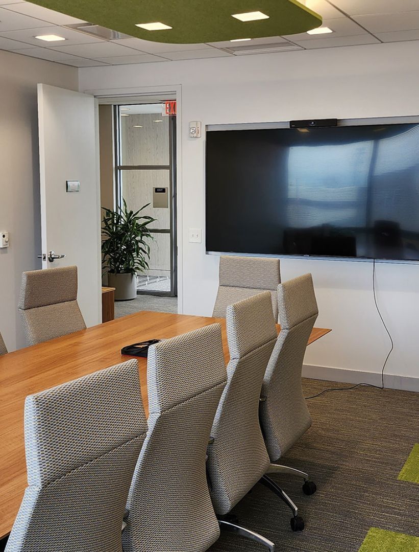 A modern conference room with a wooden table, several gray patterned office chairs, a large screen, and a green ceiling.