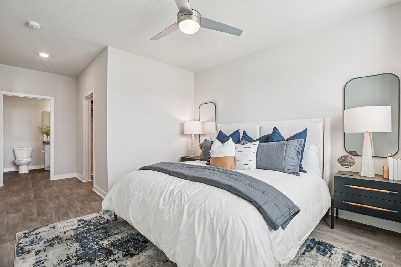 Modern apartment bedroom with a large bed, nightstands, lamps, ceiling fan, and entry to a bathroom.