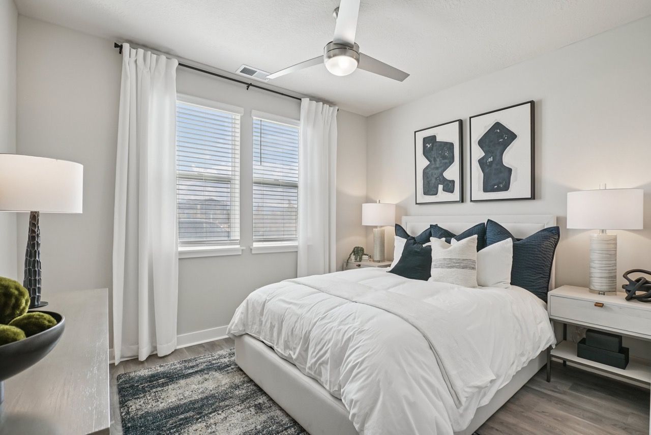 Modern apartment bedroom with white bedding, navy accent pillows, two nightstands, and a ceiling fan.