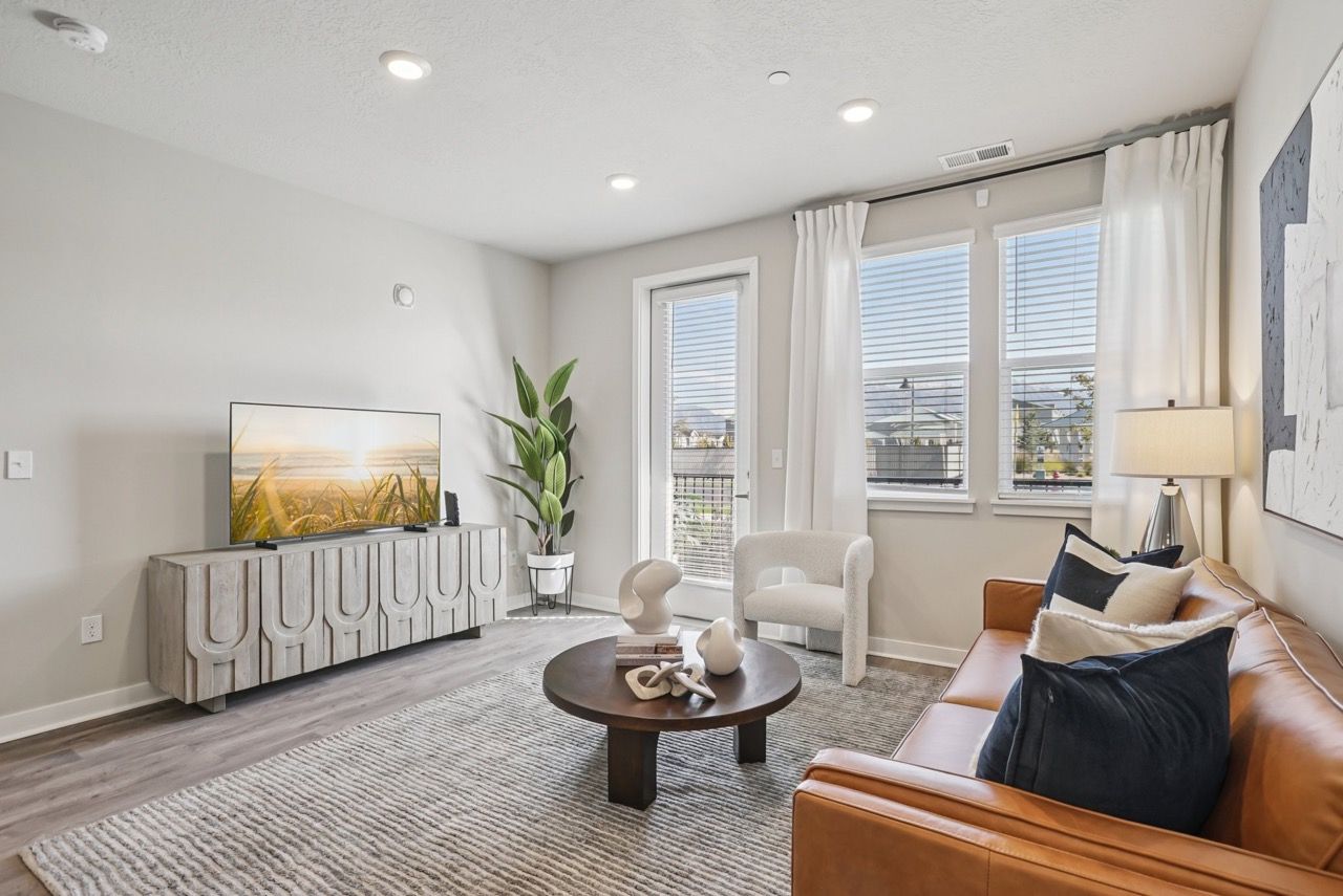 Living room in a modern apartment with a tan leather sofa, round coffee table, and large windows.