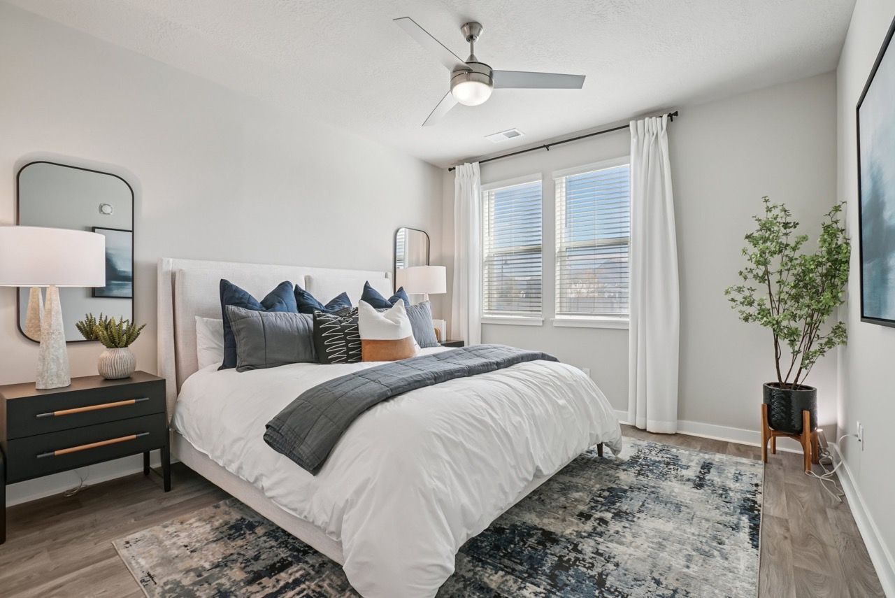 Bright bedroom with a white upholstered bed, two black nightstands, ceiling fan, and large windows with white curtains.