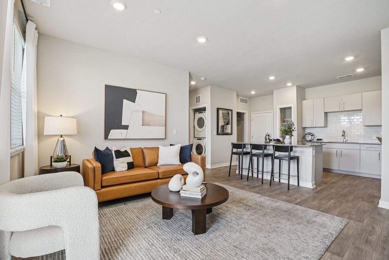 Open-concept living room with tan leather sofa, white chair, round coffee table, and a modern kitchen.