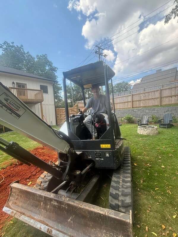 A man is driving excavation equipment in a backyard with a dog.