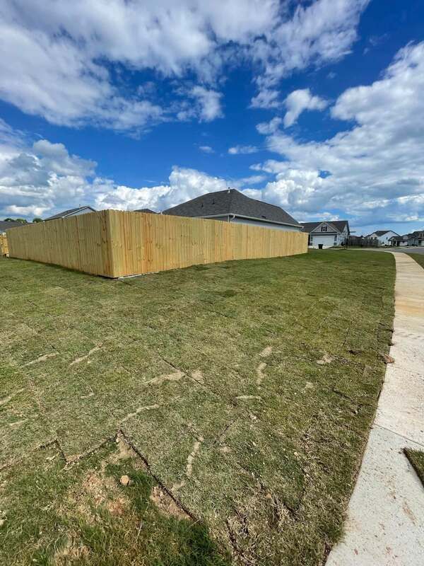 A wooden fence is in the middle of a grassy field next to a sidewalk.