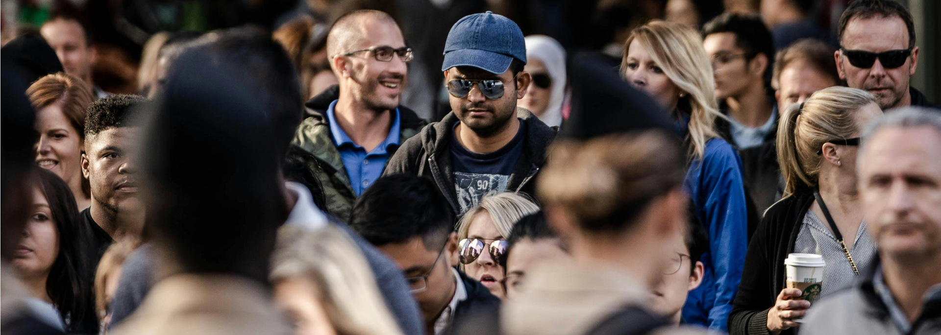 Crowd of people outdoors, with a man in a blue cap and sunglasses smiling in the center.
