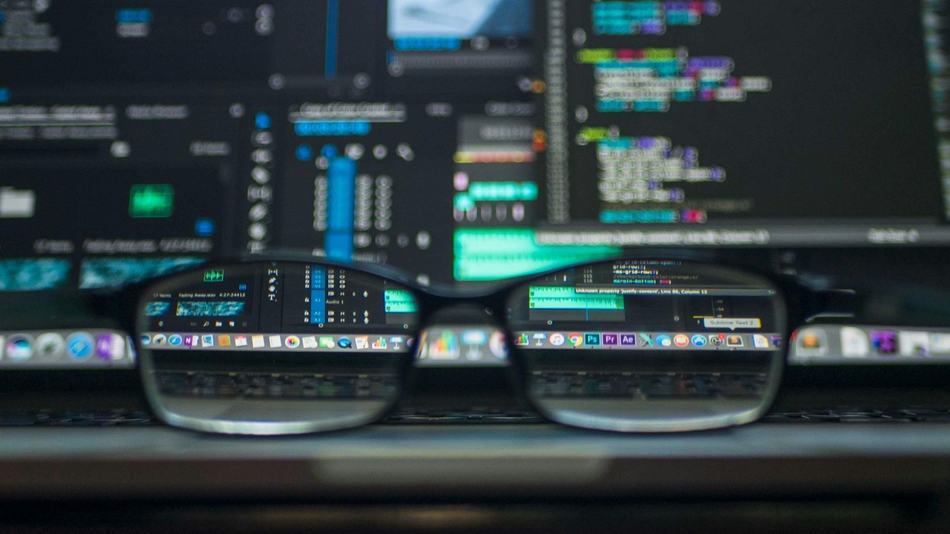 Glasses on a desk in front of a computer screen with code and colorful interface windows