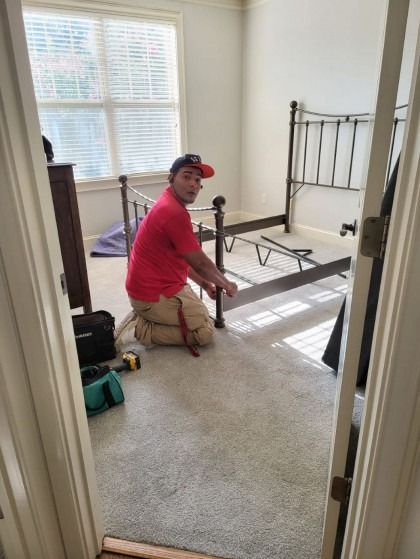 Person kneeling, assembling a metal bed frame in a room with a window, carpet, and a dresser.