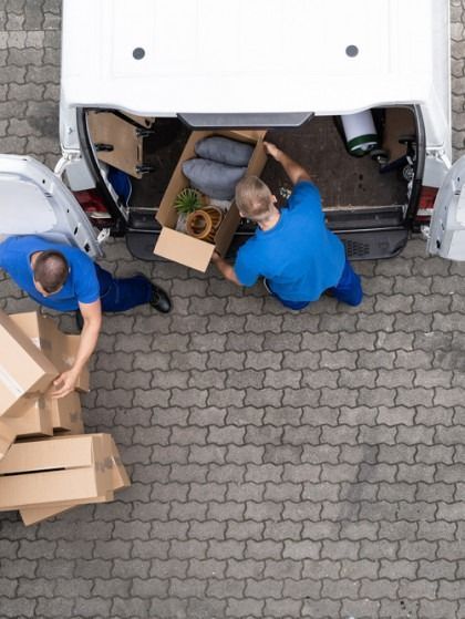 Two people in blue unload cardboard boxes from a white van parked on a brick-patterned surface.