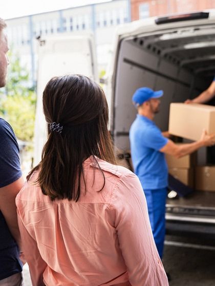Woman and man watching movers unload boxes from a white van.