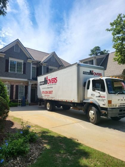 Moving truck parked in front of a gray house on a sunny day.