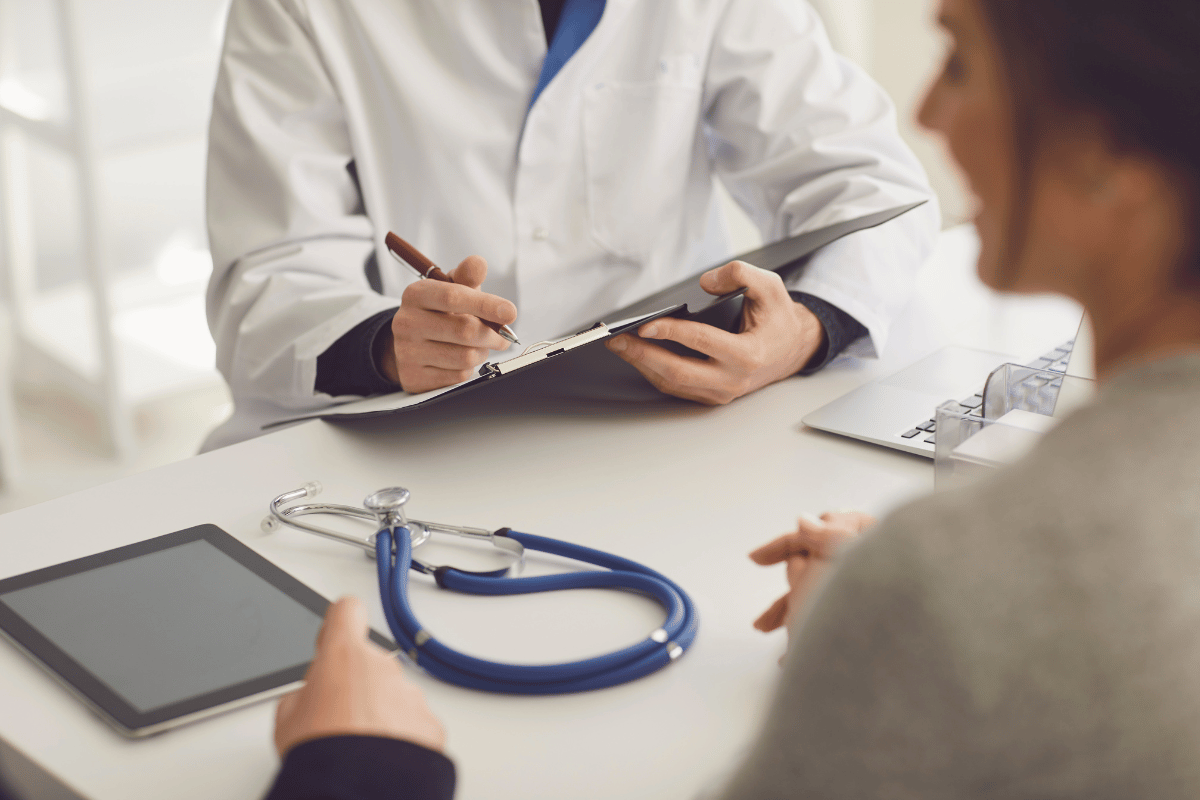 Doctor in white coat writing on clipboard; patient in foreground, stethoscope and tablet on desk.