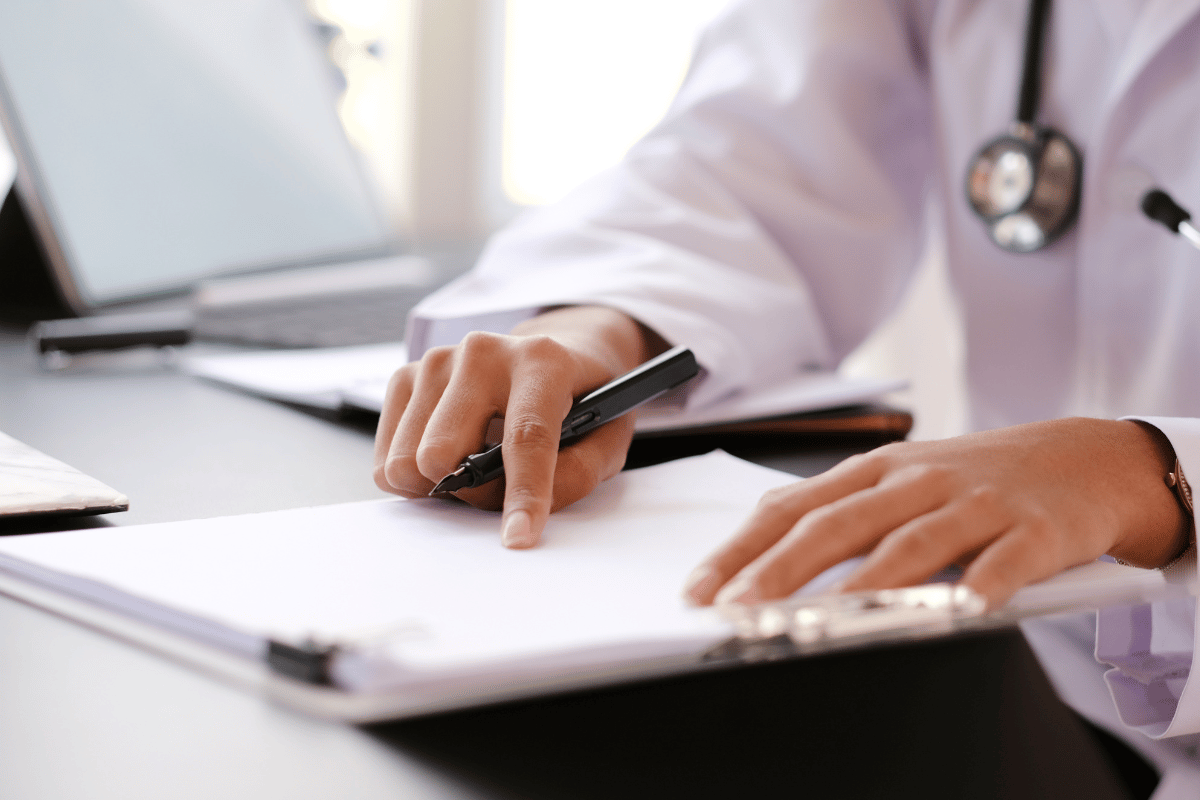Doctor in white coat writing on a clipboard at a desk, stethoscope visible.