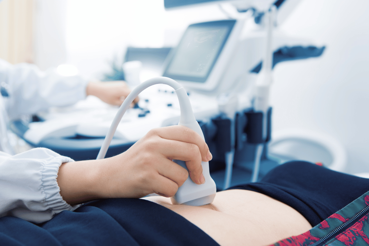 Person performing ultrasound on a patient's abdomen, using a handheld probe. Medical equipment visible in the background.